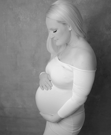 A black and white image by Meri Lee Photography captures a pregnant woman standing sideways, cradling her belly. She wears a form-fitting, off-the-shoulder gown with long hair cascading over her shoulder against a textured backdrop, celebrating the essence of motherhood.