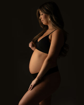 Meri Lee, a Boston South Shore photographer captures a pregnant woman standing in profile against a dark background. She wears a black bra and underwear, gently cradling her belly. Her long hair is loose, and the lighting softly highlights her form, evoking warmth and anticipation for her growing family.