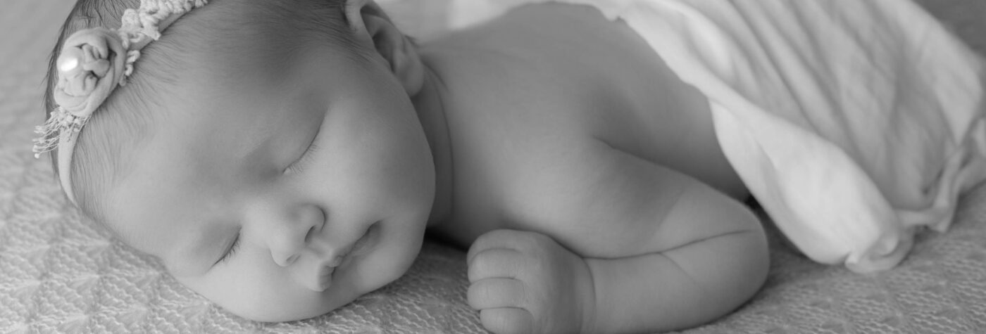 Black and white photo by Meri Lee Photography, a Boston South Shore newborn photographer captures a sleeping baby in a floral headband. Wrapped in a blanket on a textured surface, the peaceful expression and close-up angle beautifully emphasize this serene moment.
