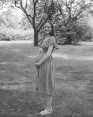 A smiling pregnant woman in a light dress stands on grass, cradling her belly. Captured by Meri Lee, a Boston South Shore photographer, she is outdoors in a park setting with trees and foliage in the background. The image, perfect for family or newborn memories, is beautifully rendered in black and white.
