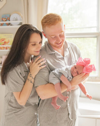 Meri Lee, a Boston South Shore photographer captures a tender moment as a couple stands in a sunlit room, both in gray pajamas. The man cradles their newborn, dressed in matching attire with a large pink bow. The woman gently touches his shoulder, smiling warmly at their baby.