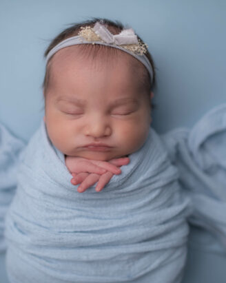 A Boston South Shore photographer captures a sleeping newborn wrapped in a light blue blanket, adorned with a delicate headband featuring a small bow and floral detail. Against a soft blue background, the baby's peaceful face and hands gently tucked under their chin convey serenity.