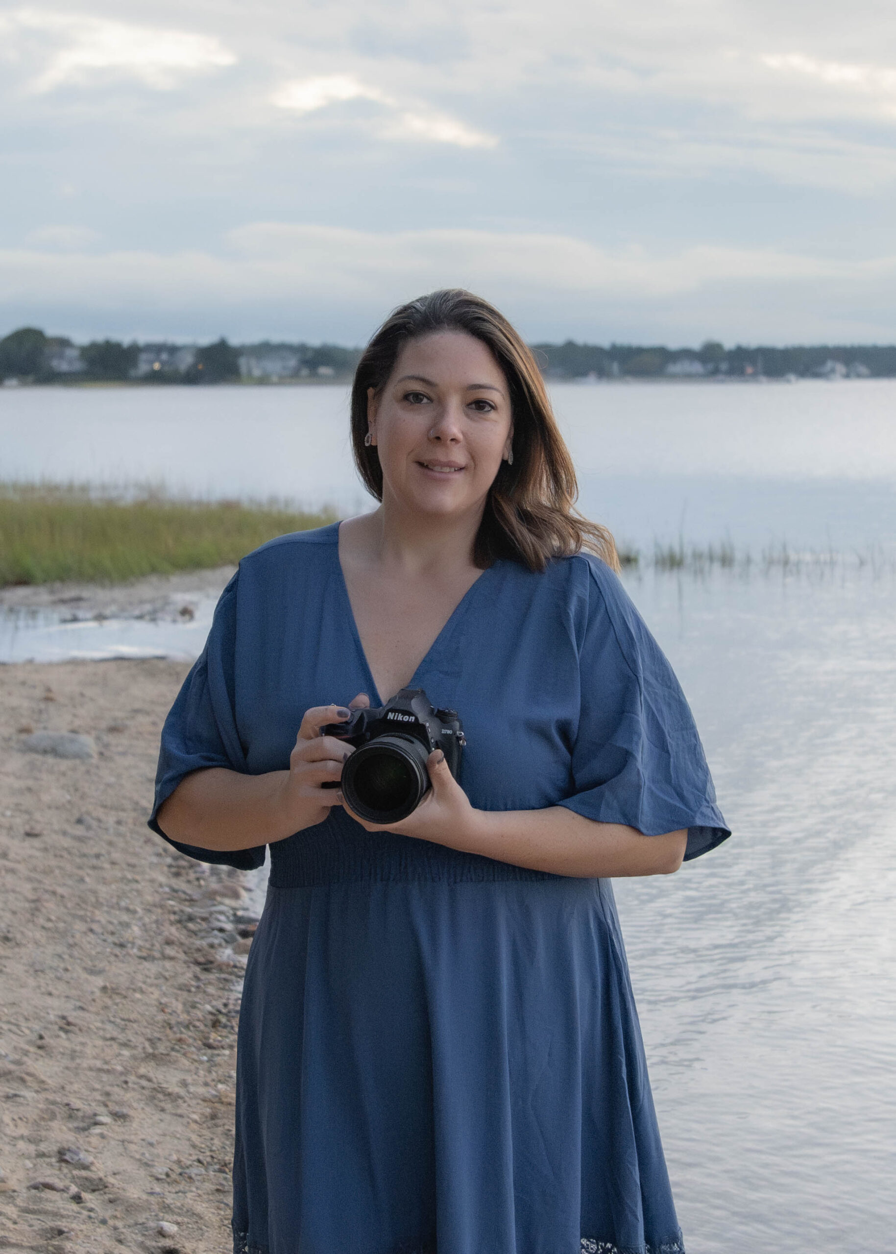 Meri, wearing a blue dress stands by a body of water during sunset, camera poised to capture the serene scene. This Boston South Shore photographer adores documenting family moments, framing each memory against the calm water and serene sky.